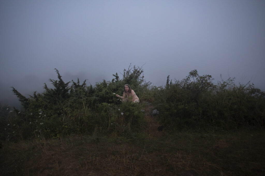 Naked woman crouching down in shrubbery, on a foggy summer morning at dawn.