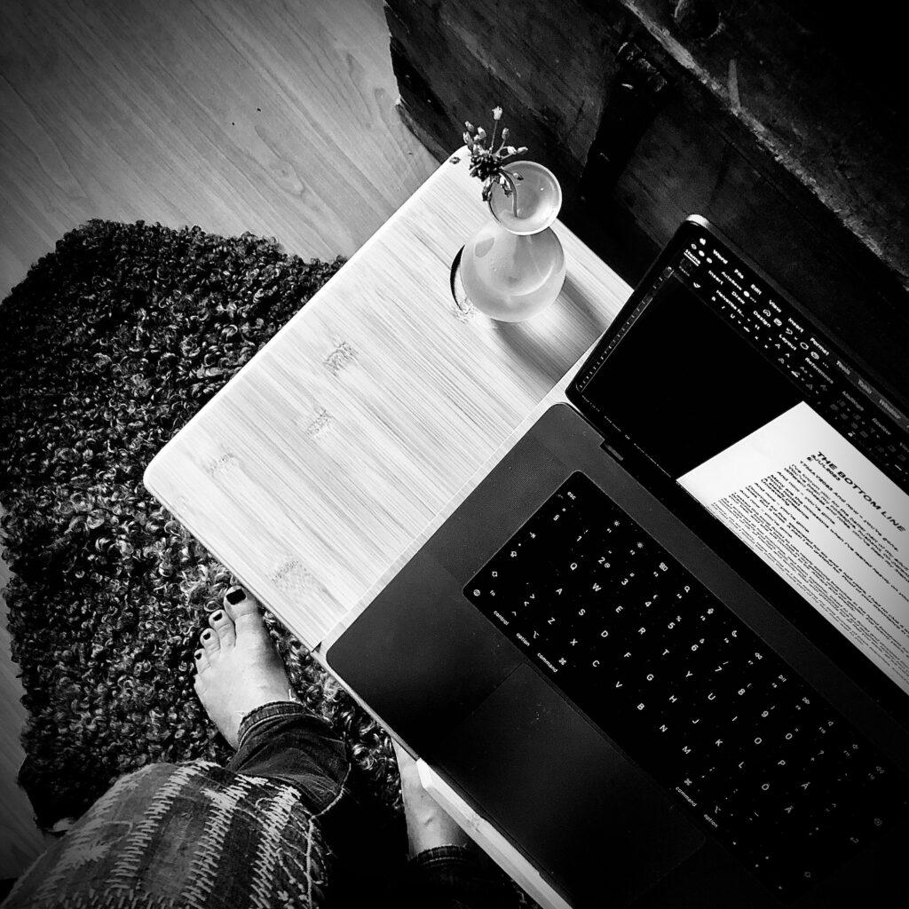 Black and white photo looking down at a computer on a small desk with a vase on it, with a pair of bare feet onto of a sheepskin lying on the floor.